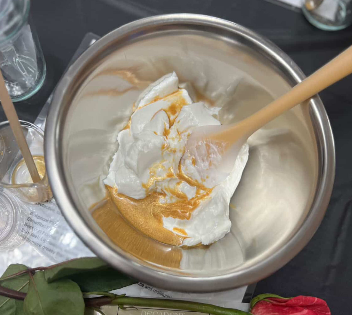 Metal bowl with the makings of a body butter and a wooden spoon on a dark surface with a red flower and paper in the background.
